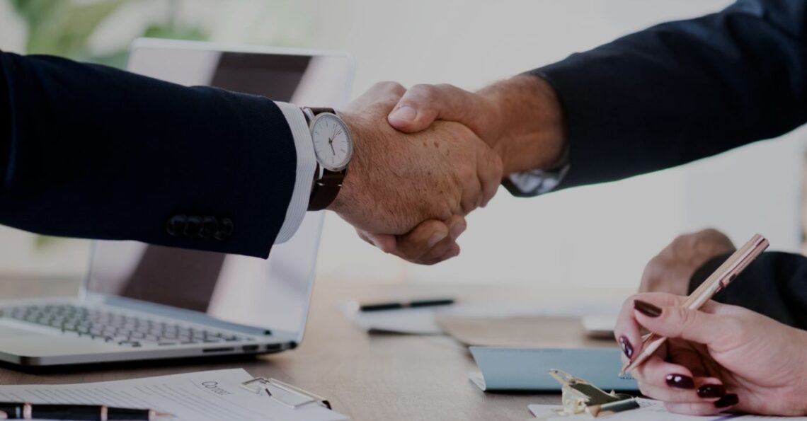 Aperto de mãos entre duas pessoas em contexto empresarial, sobre uma mesa com documentos e um portátil.