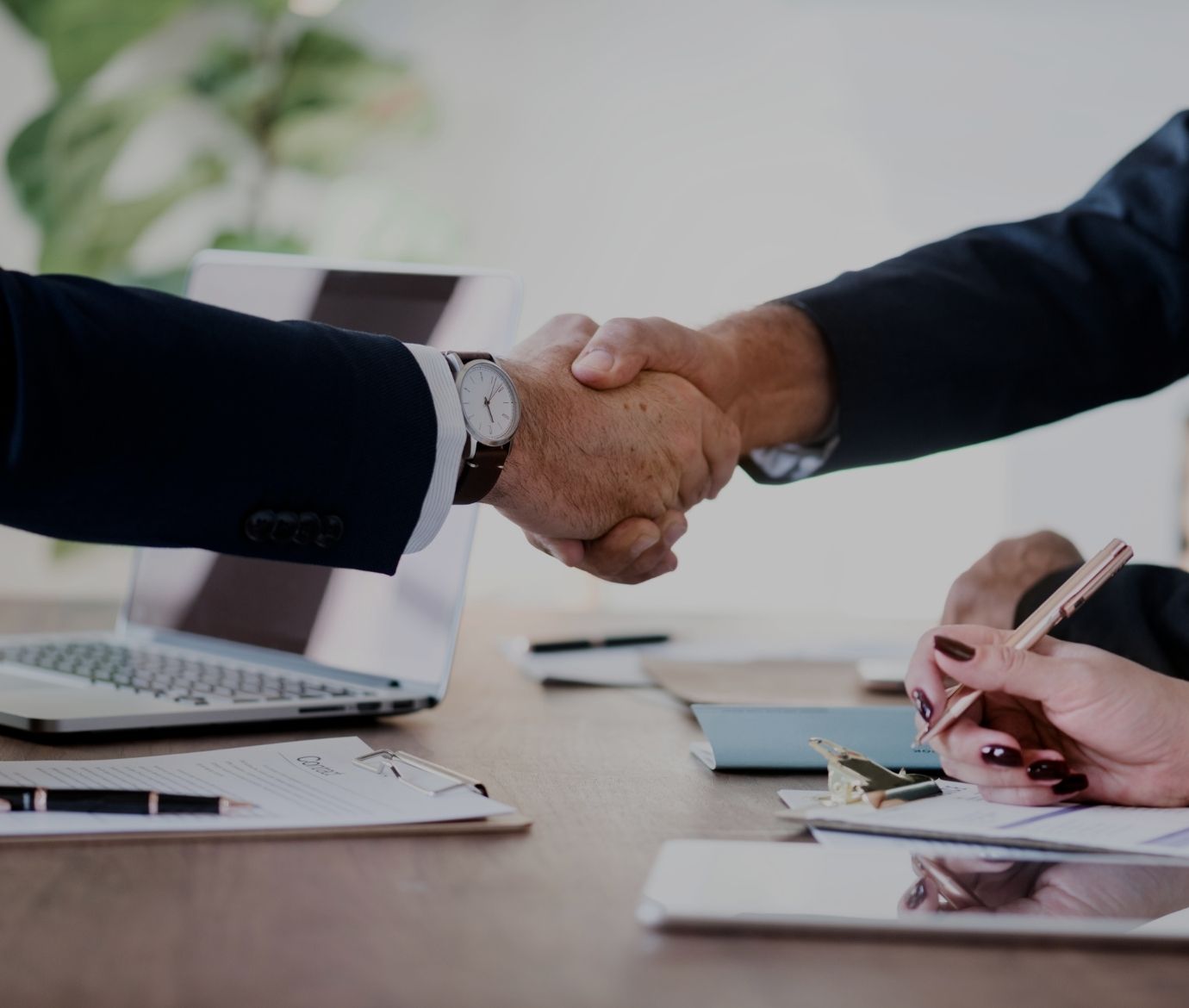 Aperto de mãos entre duas pessoas em contexto empresarial, sobre uma mesa com documentos e um portátil.