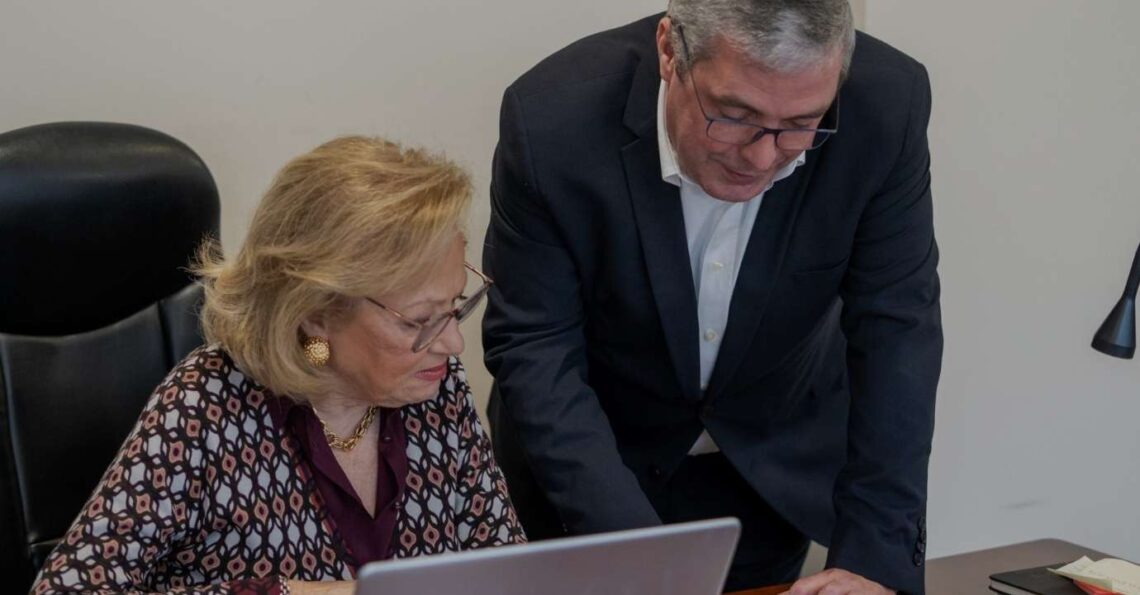 Duas pessoas em contexto profissional analisam documentos e dados num computador portátil.