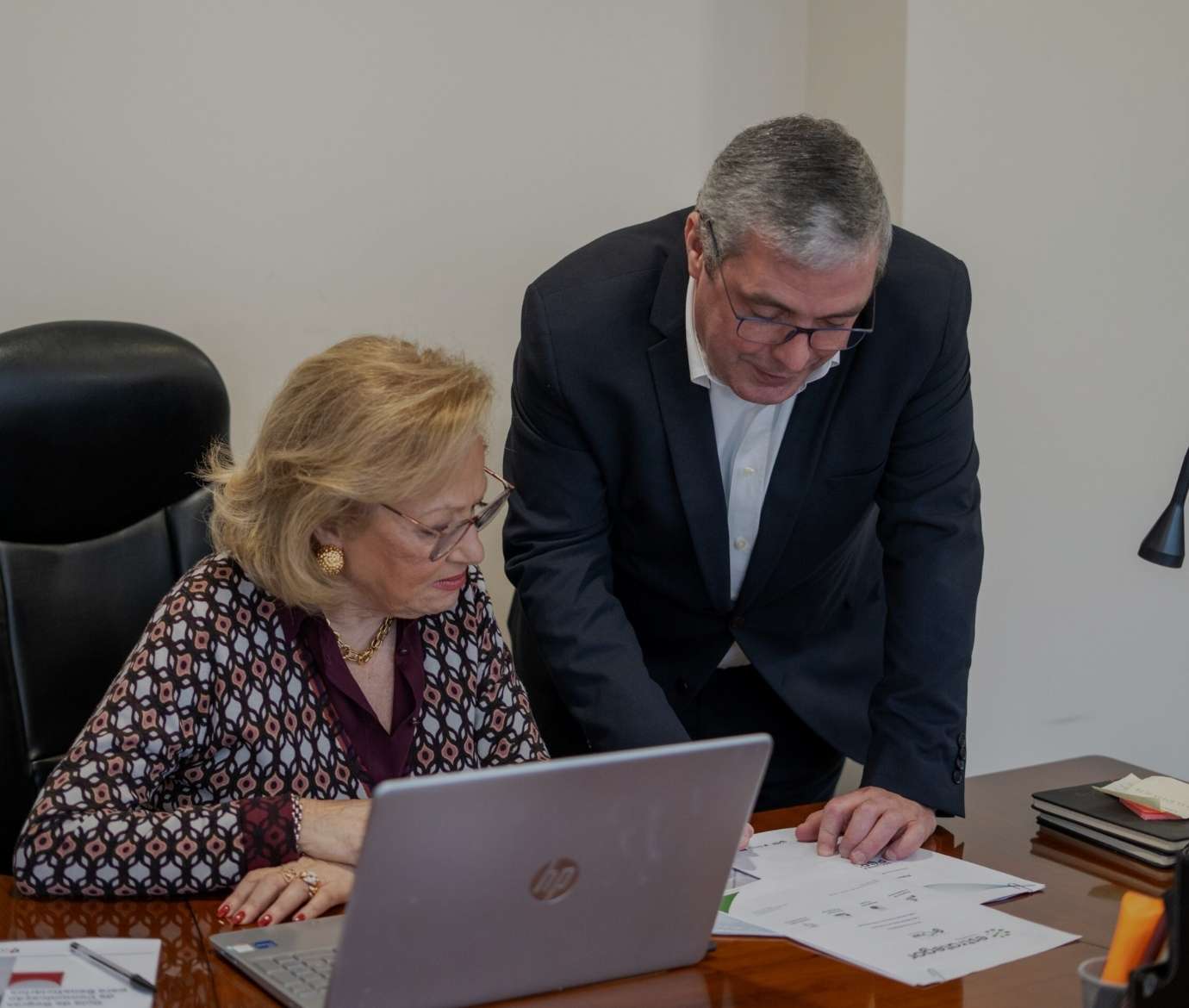 Duas pessoas em contexto profissional analisam documentos e dados num computador portátil.