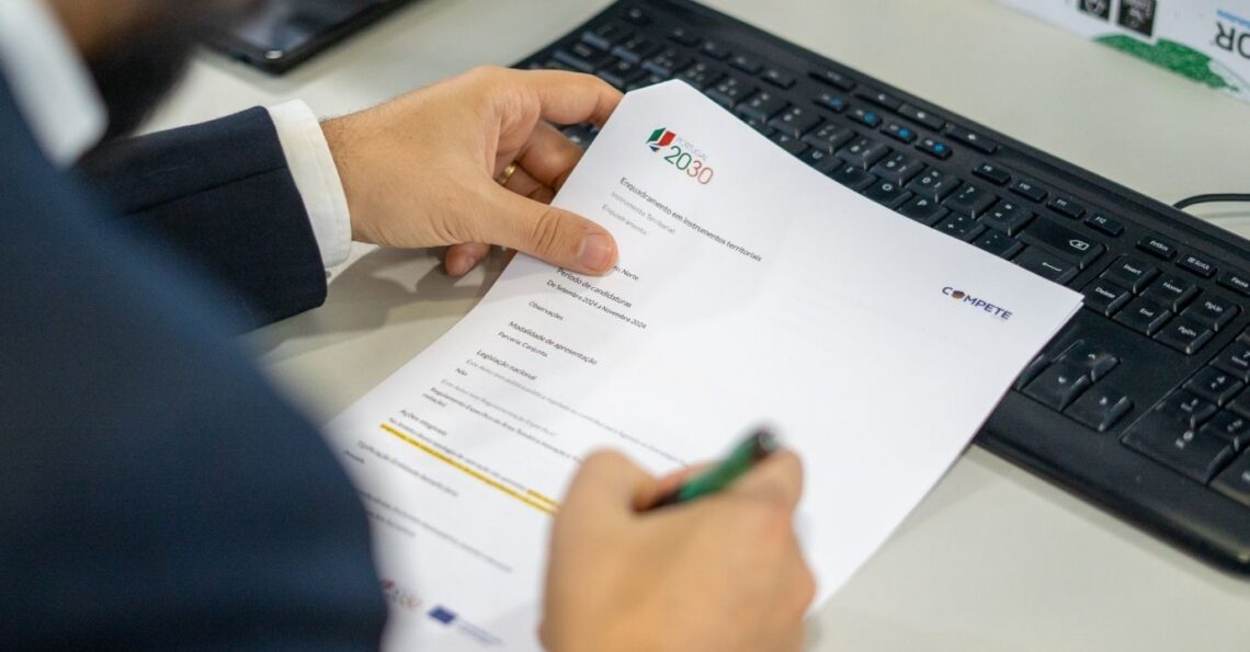 Plano aproximado das mãos de um profissional a preencher um formulário de candidatura com os logótipos "Portugal 2030" e "COMPETE" numa secretária de escritório.