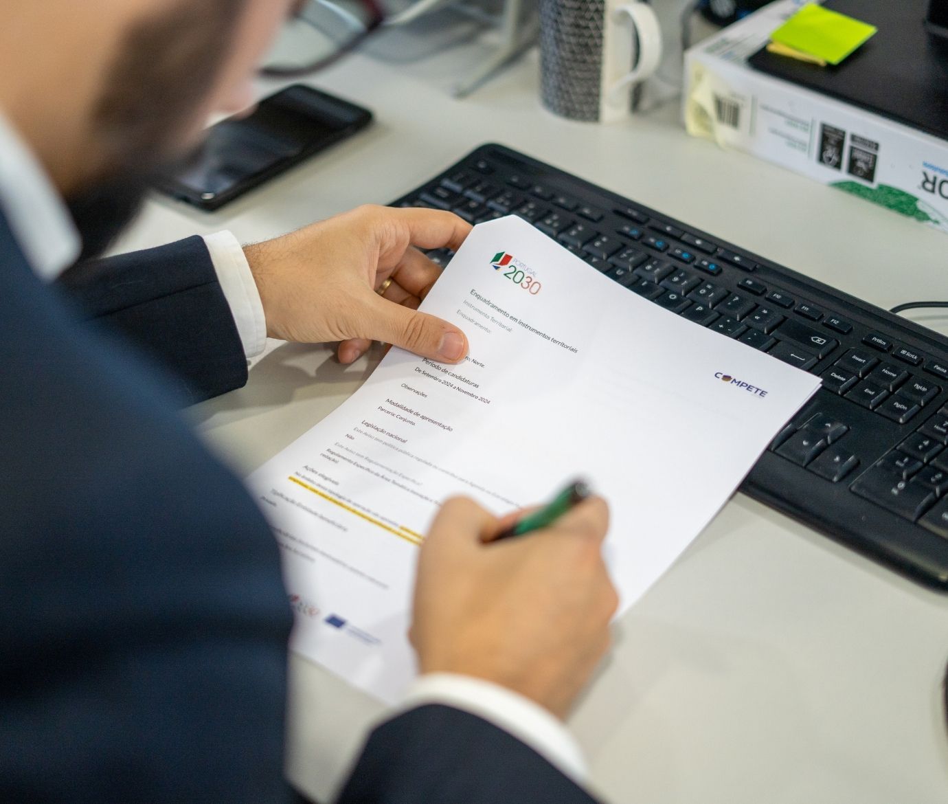 Plano aproximado das mãos de um profissional a preencher um formulário de candidatura com os logótipos "Portugal 2030" e "COMPETE" numa secretária de escritório.
