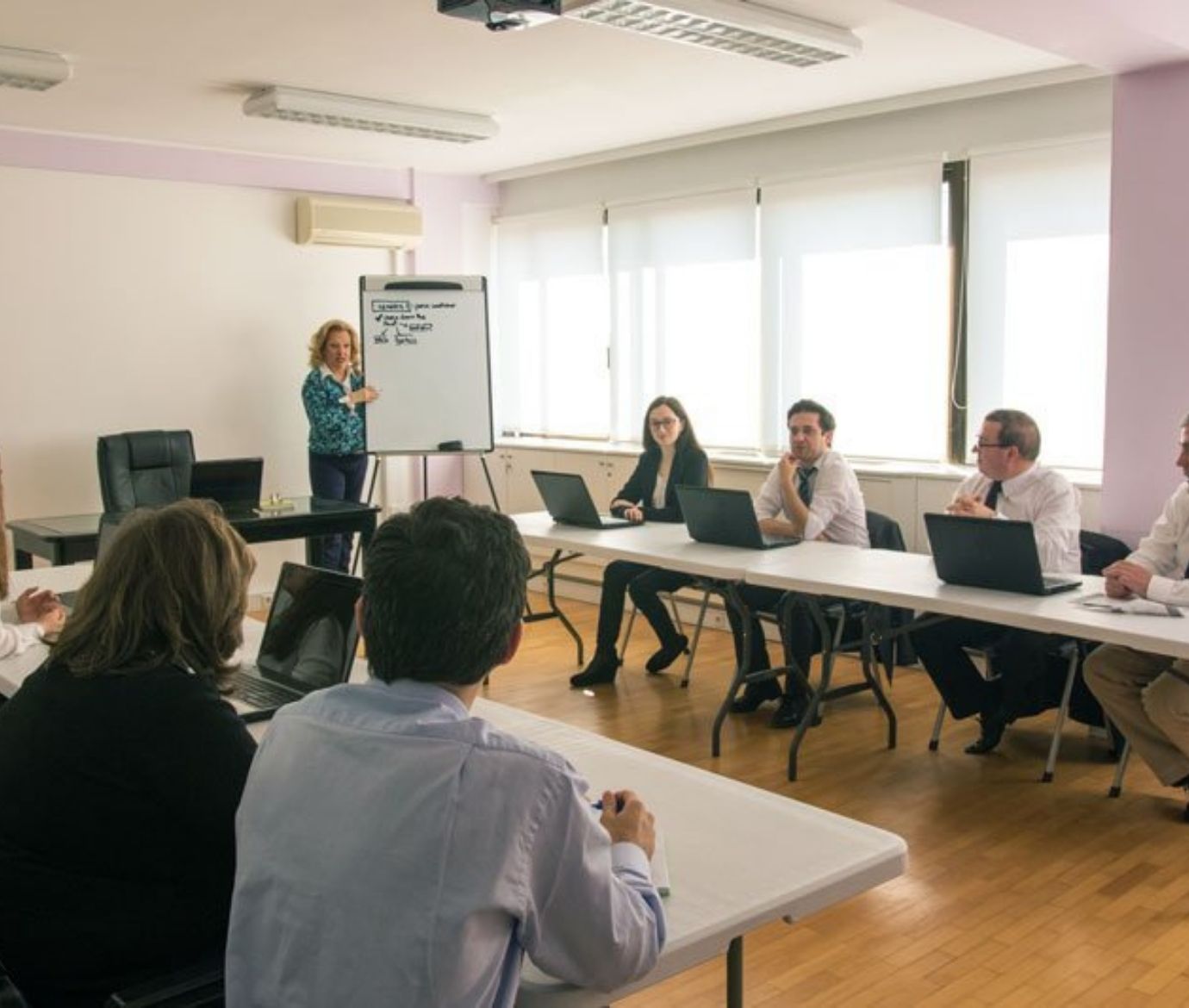 Sessão de formação corporativa onde uma formadora apresenta conteúdos num flipchart para um grupo de profissionais sentados em forma de U com computadores portáteis.