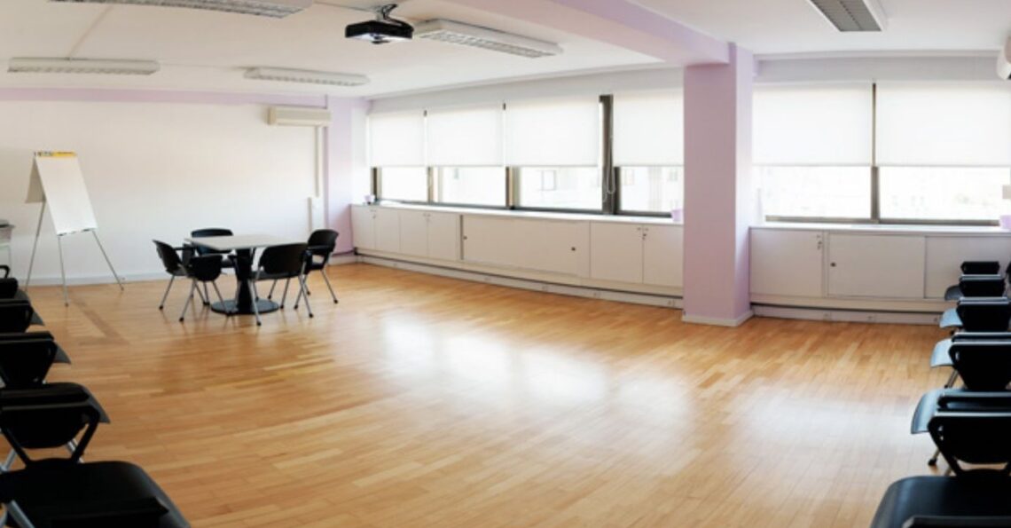 Vista panorâmica de uma sala de formação vazia e espaçosa, com chão de madeira, muita luz natural e cadeiras organizadas em círculo e filas.