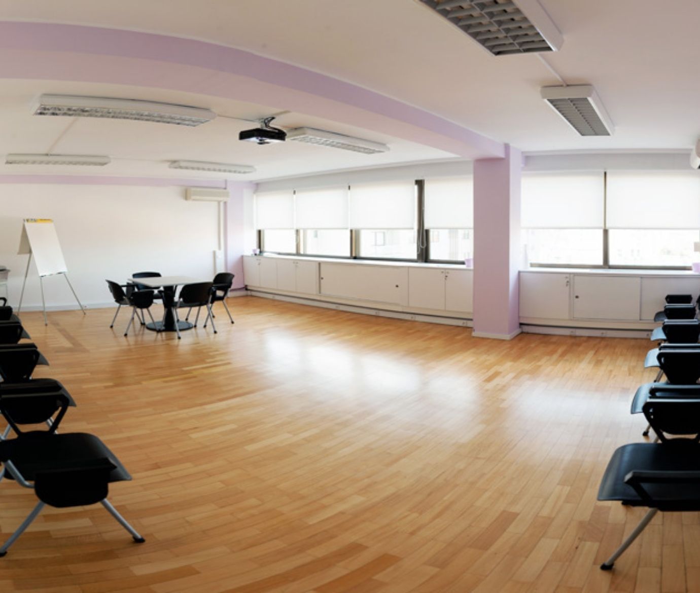 Vista panorâmica de uma sala de formação vazia e espaçosa, com chão de madeira, muita luz natural e cadeiras organizadas em círculo e filas.