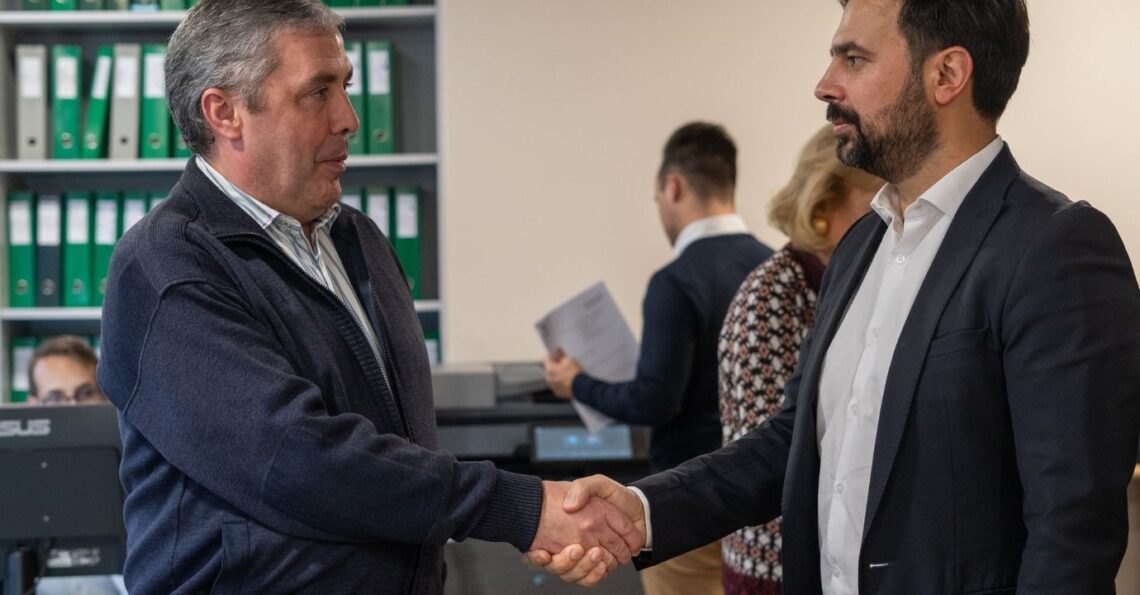 Dois homens de negócios trocam um aperto de mão formal num escritório, simbolizando um acordo ou parceria, com estantes de arquivos em segundo plano.