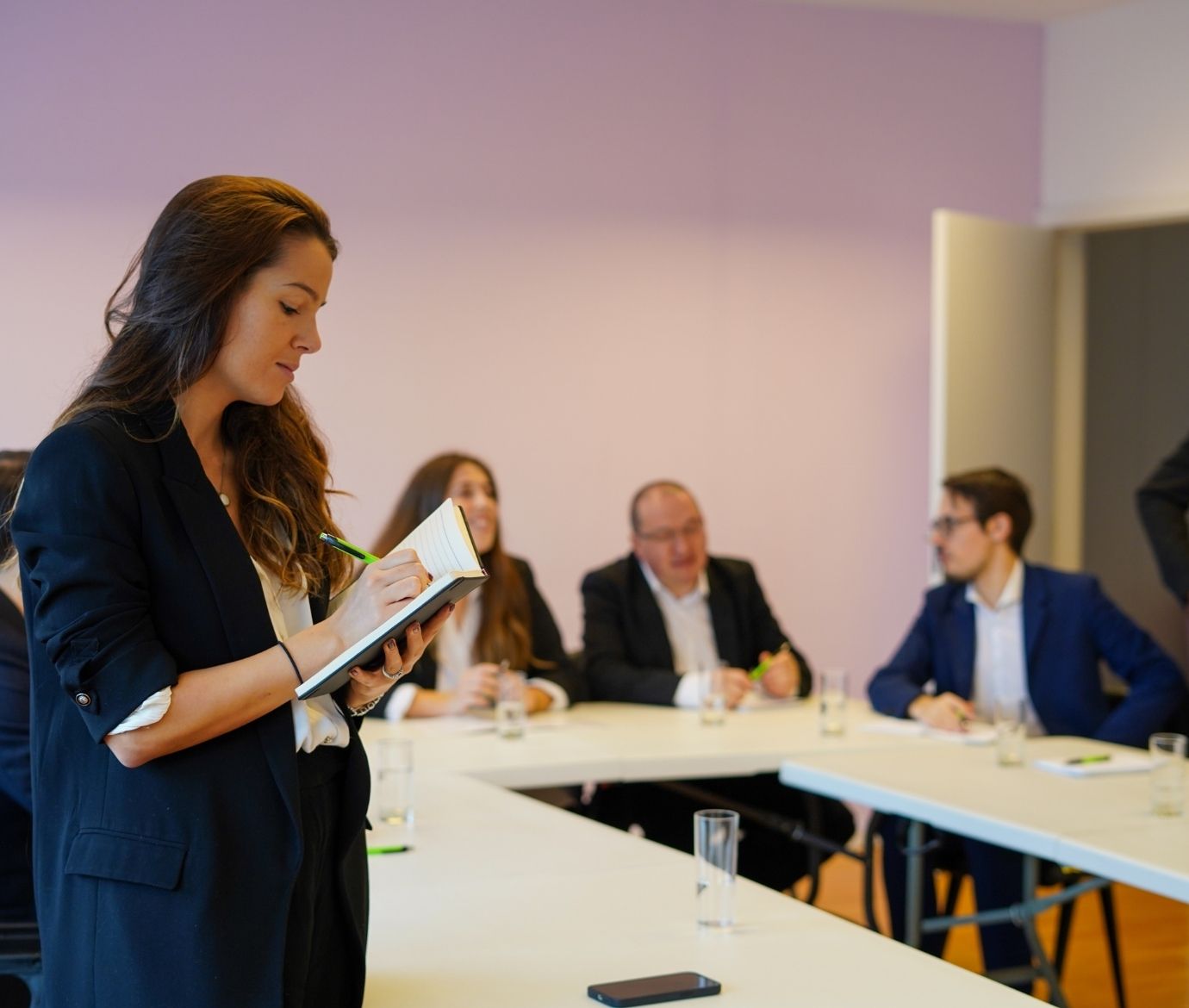 Uma mulher em primeiro plano escreve num bloco de notas durante um workshop, com outros participantes desfocados sentados em mesas ao fundo.