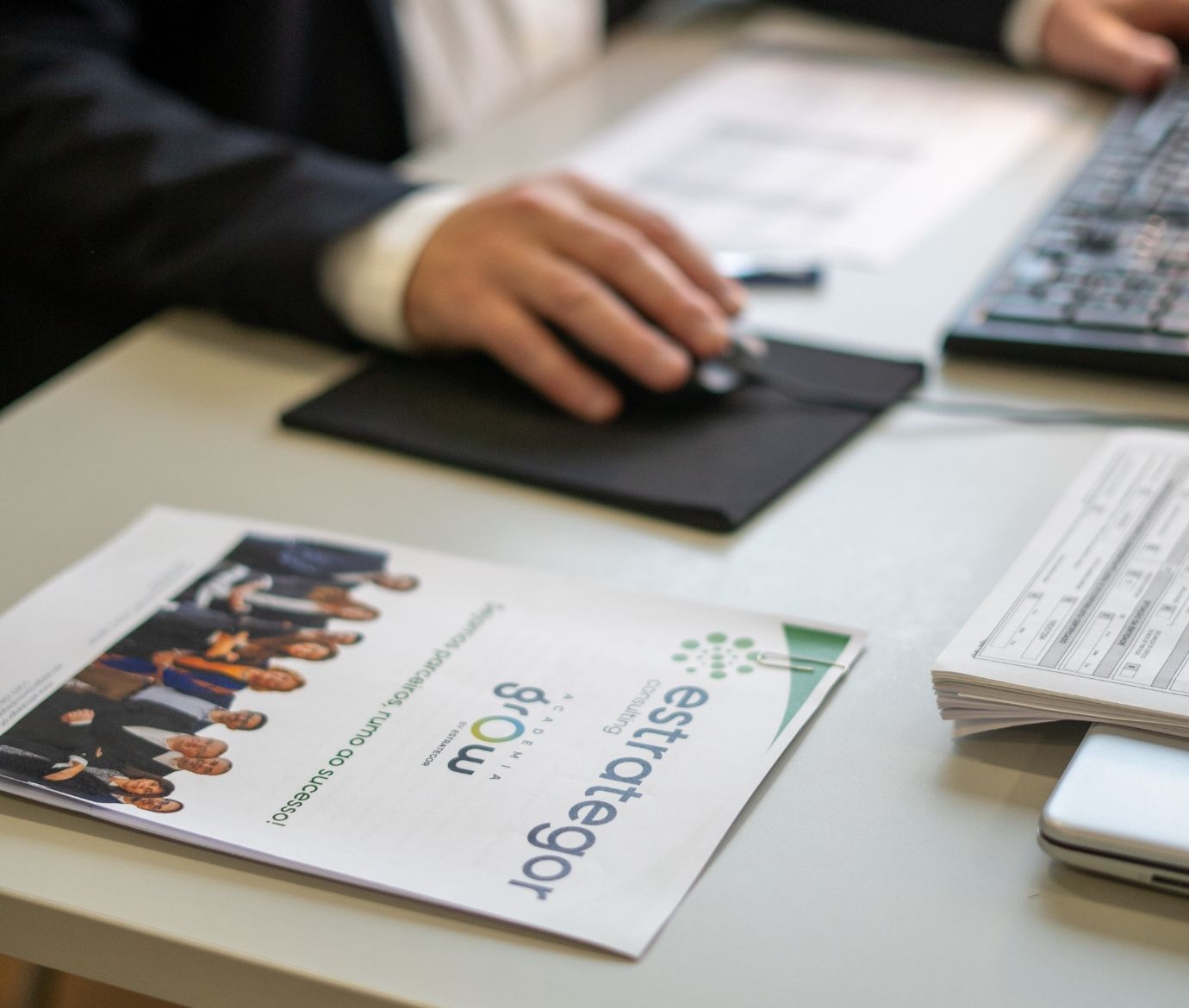 Foco seletivo num folheto da "Estrategor" e "Academia Grow" sobre uma mesa, com a mão de um utilizador a usar um rato de computador em fundo desfocado.